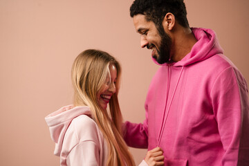 Wall Mural - Romantic fashion studio portrait of a biracial young couple in love in hoodie posing over pink background.