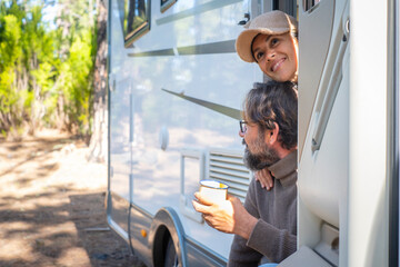 Young couple of tourists with camper motor home sitting at the door enjoing nature outdoors leisure feeling. Travel people and mountains destination concept. Man and woman living van life together