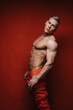 Sexy blond in red sweatpants with  curved back. Muscular man in sportswear on  red background. Handsome guy with six pack abs taking off his pants in studio.