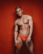 © Serge Lee - Young sexy muscular man in red swimwear with smartphone in his hand. Shirtless male model in red speedo at red background. Cover shot with blond handsome guy in studio.