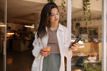 Calm young caucasian woman is using smartphone enjoying drink standing in doorway of cafe. Brunette wears t-shirt, pants and shirt. Lifestyle concept