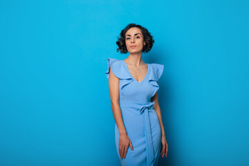 Wall Mural - Close-up photo of a charming healthy woman in a blue dress, who is looking in the camera and smiling, while standing and posing isolated on blue background