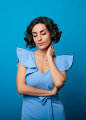 Wall Mural - Vertical photo of a gorgeous tender woman in a elegant blue dress, who is looking down and while standing and posing isolated on blue background