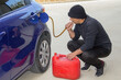 Image of a thief dressed all in black sucking gasoline from a parked car with a rubber hose. Reference to the current expensive fuel.