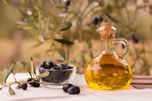 Olive Oil In A Glass Bottle And Green Olives On The Background Of Olive Branches In The Garden