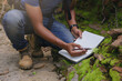 © Panumas - Crop image of Biologist or botanist recording information about small tropical plants in forest. The concept of hiking to study and research botanical gardens by searching for information.