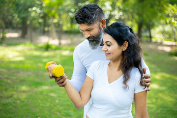 Indian trainer helps a beautiful woman with dumbbell workout at park, working on arms strength, Asian athletic lady exercising outdoor with coach in nature, fitness concept.