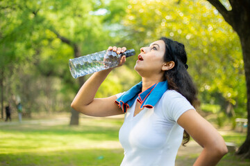 Young tired thirsty indian woman drinking water in summers at park. Daily workout, hydrate concept. Sports girl. 