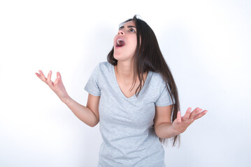 Wall Mural - young beautiful brunette woman wearing grey t-shirt over white wall crazy and mad shouting and yelling with aggressive expression and arms raised. Frustration concept.