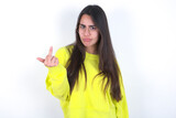 Young beautiful brunette woman wearing yellow hoodie over white wall shows middle finger bad sign asks not to bother. Provocation and rude attitude.