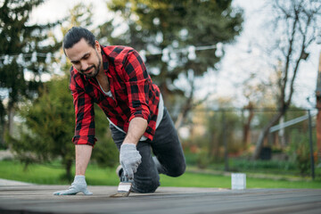 A young man in home clothes paints a wooden deck in the garden with a brush. The householder varnishes or protects the boards of the campfire site with protective oil.