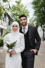 Wall Mural - happy groom posing with hand in pocket near muslim bride in hijab with wedding bouquet.