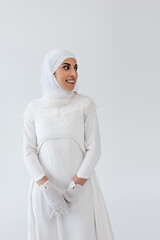 Wall Mural - joyful muslim woman in hijab and wedding dress isolated on grey.