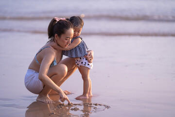 mom with baby girl on the beach by write shape heart on sand in evening time with sunset light