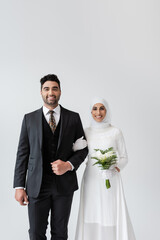 Wall Mural - happy muslim bride with bouquet standing near cheerful groom isolated on grey.