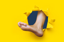 A Left Woman's Foot Appears In A Torn Hole In Yellow Paper. Healthy Legs Concept. Joke. Background With Copy Space.