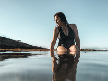 Woman In Full Piece Bathing Suit Sitting On Reef Ledge Looking Into Glassy Ocean Water