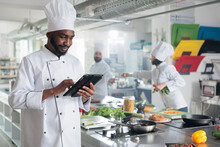 Gastronomy Expert With Handheld Device Standing In Restaurant Professional Kitchen While Looking For Dinner Recipes. Head Cook With Tablet Brainstorming Garnish Ideas For Gourmet Cuisine Meal.