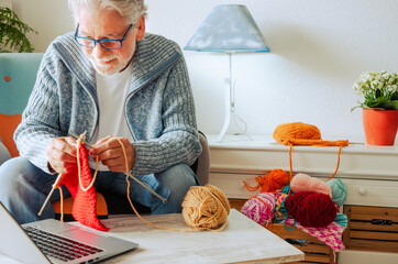 Wall Mural - Senior man at home follow online knit tutorial to relax and enjoy resting home leisure activity sitting on the sofa - male people knitting with computer class help - people inner lifestyle hobby