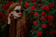 Beautiful fashionable redhead freckled woman wearing trendy black cat eye sunglasses, pearl hoop earrings, posing in red rose garden. Copy, empty space for text