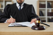 © Studio Romantic - Close up of a gavel on a wooden table in the courtroom, with a male judge putting his signature on some legal documents in the background. Court trial, justice, legislation, law enforcement concepts