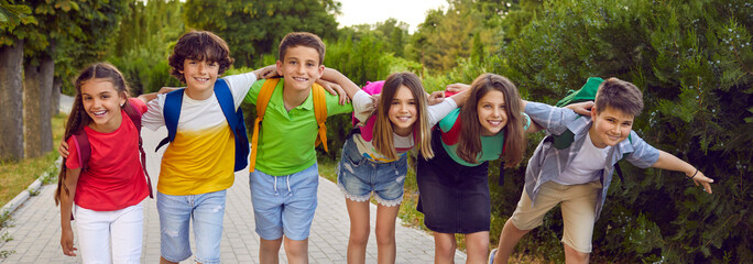 Joyful elementary school boys and girls posing in kids clothes on joint summer walk in park, symbolizing happy childhood full of positive emotions, hugging each other standing on path. Panoramic shot