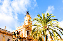 Clock Tower Building. Large Clock Tower With Hour Hands That Shows Time. Palm Trees On The Background Of The Facade Of A Residential Building. Architecture Of Modern Buildings In Valencia, Spain.