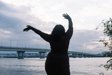 Rearview of curly multicultural woman silhouette looking at the water horizon, sea beach and cloudy blue sky. Dancing, gesturing by raising arms in air. Vacation, relaxation in summer. Body positive