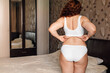 © Юля Бурмистрова - Curly plus size woman in white underwear standing near bed in bedroom and pinching sagging folds on back. Fat burning treatment of overweight body. Weight loss program, anticellulite massage.