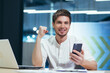 © Liubomir - Portrait of a young businessman man smiles and rejoices in victory holds in his hands the phone looks at the camera, holds his hand up gesture of victory and triumph