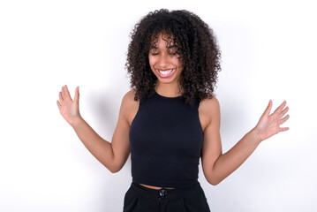 Emotive Young beautiful girl with afro hairstyle wearing black tank top over white background laughs loudly, hears funny joke or story, raises palms with satisfaction, being overjoyed amused by friend