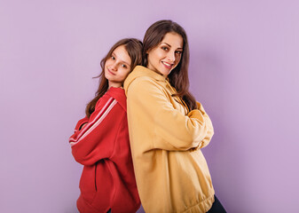 Wall Mural - Happy smiling fun young mother and cute daughter posing back to back in fashion trendy hoodie together on purple studio background with empty copy space. Happy loving family