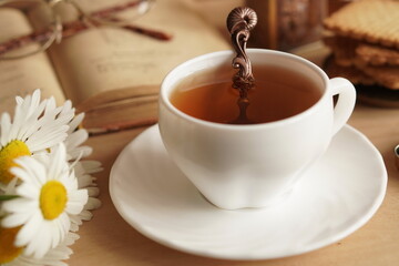 a composition of a white porcelain cup with tea on a saucer and a decorative spoon with a bouquet of white chamomile flowers, a book with metal-rimmed glasses. The concept of a summer breakfast.