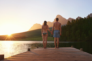 Wall Mural - Time for a sunset dip. Rearview shot of a young couple in swimsuits standing on a dock at sunset.