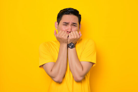 Worried young Asian man in casual t-shirt biting nails, reacting to scary news isolated on yellow background. People lifestyle concept