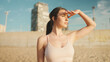 © Andrii Nekrasov - Young athletic woman with long ponytail wearing beige sports top walks along the beach at dawn on modern city background