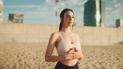 Young athletic woman with long ponytail wearing beige sports top is doing breathing exercises in the rising sun