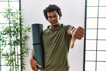 Canvas Print - African man with curly hair holding yoga mat at studio looking unhappy and angry showing rejection and negative with thumbs down gesture. bad expression.