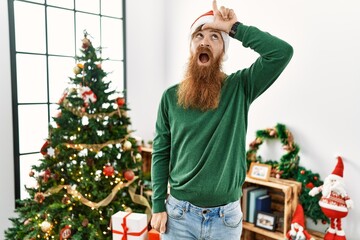 Wall Mural - Redhead man with long beard wearing christmas hat by christmas tree making fun of people with fingers on forehead doing loser gesture mocking and insulting.