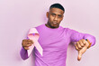 Young black man holding pink cancer ribbon with angry face, negative sign showing dislike with thumbs down, rejection concept