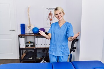 Wall Mural - Young physiotherapist woman working at pain recovery clinic smiling cheerful offering hands giving assistance and acceptance.