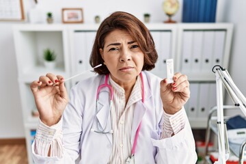 Poster - Middle age hispanic doctor woman holding coronavirus infection nasal test clueless and confused expression. doubt concept.