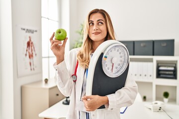 Poster - Young blonde doctor woman holding weighing machine and green apple smiling looking to the side and staring away thinking.