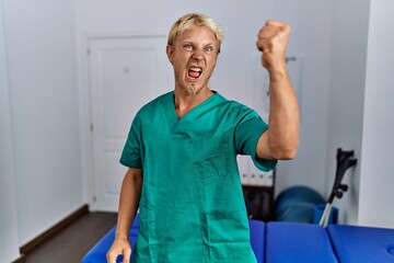 Poster - Young blond man wearing physiotherapist uniform standing at clinic angry and mad raising fist frustrated and furious while shouting with anger. rage and aggressive concept.