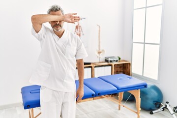 Poster - Middle age hispanic therapist man working at pain recovery clinic covering eyes with arm, looking serious and sad. sightless, hiding and rejection concept