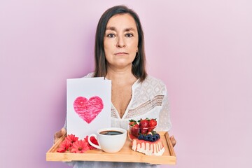 Wall Mural - Middle age hispanic woman holding tray with breakfast food and heart draw relaxed with serious expression on face. simple and natural looking at the camera.