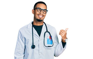 Young african american man wearing doctor uniform and stethoscope smiling with happy face looking and pointing to the side with thumb up.