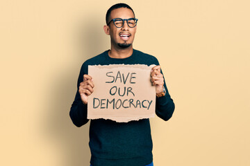 Young african american man holding save our democracy protest banner winking looking at the camera with sexy expression, cheerful and happy face.