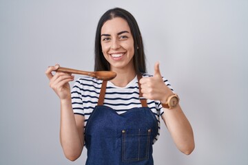 Poster - Young brunette woman wearing apron tasting food holding wooden spoon smiling happy and positive, thumb up doing excellent and approval sign