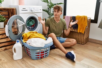 Sticker - Young caucasian man putting dirty laundry into washing machine clueless and confused expression with arms and hands raised. doubt concept.